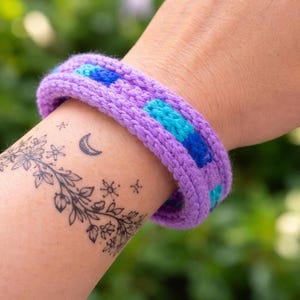 May include: A knitted bracelet in shades of purple, blue, and turquoise. The bracelet is a thick band with a textured appearance. The design features a repeating pattern of blue and turquoise squares against a purple background. The bracelet is approximately 2 inches wide.