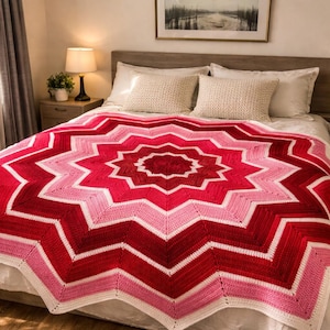 Peut inclure: Une couverture au crochet faite à la main en forme d'étoile, dans des tons de rouge et de rose, avec une bordure blanche. La couverture est présentée sur un lit avec une literie blanche. Le motif présente un cercle central rouge plus foncé qui rayonne vers l'extérieur.