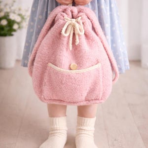 May include: A soft pink knitted backpack with a drawstring closure and a front pocket. The backpack has a cream-colored pocket trim and a wooden button. The bag is held by a person wearing a blue polka dot dress and white socks.