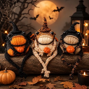 Peut inclure: Trois décorations de grenouilles d'Halloween faites à la main, chacune tenant une petite citrouille. Les grenouilles sont tricotées en noir, orange et crème, et portent des chapeaux de sorcière. Elles sont disposées sur une bûche de bois avec une pleine lune et des chauves-souris en arrière-plan.