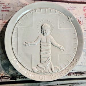 May include: A round, off-white ceramic plate featuring a central embossed figure of a child with arms outstretched, positioned in front of a cross. The text 19-KING OF KINGS-70 is embossed along the bottom rim, with a star in the upper right.