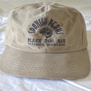 May include: Tan baseball cap with a curved brim. The cap features the text "Spanish Peaks" above a black dog graphic, and "Black Dog Ale Bozeman, Montana" below. The cap is made of a durable fabric and has a classic, casual style.