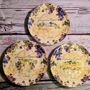 May include: Three round ceramic plates featuring a cream-colored background with a decorative border of purple and blue grapes. Each plate displays a central illustration of a vineyard landscape and the text Francesco Vigneto Vino di Italia.