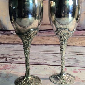 May include: Two silver-toned metal goblets featuring smooth, reflective bowls and textured stems designed with intricate, raised grape vine and leaf motifs that extend down to the circular bases, creating a detailed, ornate appearance.