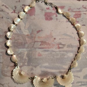 May include: A necklace featuring a strand of iridescent, crescent-shaped shell beads. The bottom section displays three larger, fan-shaped carved shell pendants with intricate line detailing. The components are connected by metal links and a hook clasp.