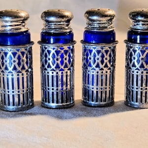 May include: Four cylindrical salt and pepper shakers featuring deep cobalt blue glass inserts encased in intricate silver-toned metal filigree sleeves with matching rounded silver-toned metal caps with small holes at the top for dispensing.