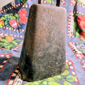 May include: A weathered, trapezoidal metal cowbell with a dark, rusted patina and a rectangular handle at the top. The surface shows significant oxidation and texture, with a deep brown and black color palette throughout the entire object.