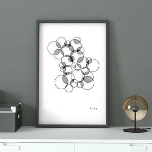 Könnte beinhalten: Ein gerahmter Kunstdruck mit einem abstrakten Schwarz-Weiß-Design aus sich überlappenden Kreisen. Das Kunstwerk wird auf einer weißen Oberfläche zusammen mit einer kleinen Holzbox, einer grauen Aufbewahrungsbox und einer dekorativen Lampe präsentiert.