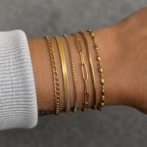 May include: A set of six gold-toned bracelets worn together on a wrist. The collection includes a classic curb chain, a flat herringbone chain, a thin box chain, a long oval paperclip link chain, a fine snake chain, and a beaded chain design.