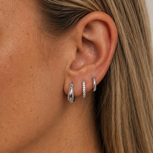 May include: Three silver hoop earrings are displayed on an ear. The first is a thick, polished silver hoop. The second is a smaller hoop with a row of small, clear stones. The third is a thin, polished silver hoop.