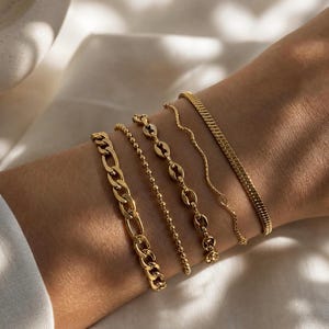 May include: Five gold bracelets of varying designs are displayed on a wrist. The bracelets include a chain link, a beaded chain, a chain with oval links, a wavy chain, and a flat, textured chain. The jewelry is set against a soft, neutral background.