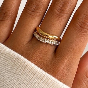 May include: A stack of two gold rings worn on a finger. The top ring is a smooth, polished gold band with a slightly wavy, organic shape. The bottom ring is a gold band encrusted with a single row of small, clear, sparkling round-cut stones.