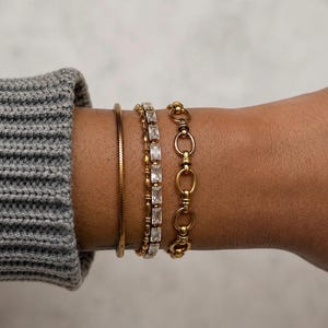 May include: A set of three gold-toned bracelets worn together. The first is a thin, flat snake chain. The second is a tennis-style bracelet featuring rectangular clear stones. The third is a chain composed of interlocking oval and round links.