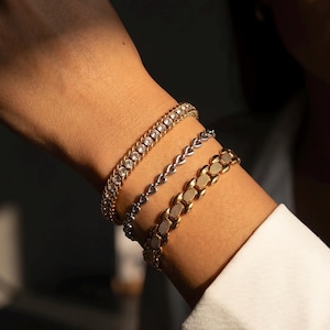 May include: Three bracelets on a wrist. The top bracelet is gold-toned with clear stones. The middle bracelet is silver-toned with heart-shaped links. The bottom bracelet is gold-toned with a textured pattern. The person is wearing a white shirt.