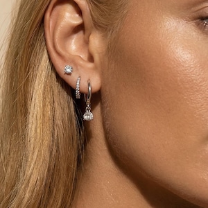 May include: A close-up of an ear adorned with three pieces of jewelry. A small, round stud earring is in the upper lobe. A small hoop earring with a row of stones and a dangling teardrop-shaped earring complete the set.