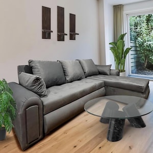 Puede incluir: Un sofá seccional gris con múltiples cojines y una chaise longue. Una mesa de centro redonda de cristal con base de mármol negro se encuentra frente al sofá. Tres estantes de pared de madera oscura están montados encima del sofá.