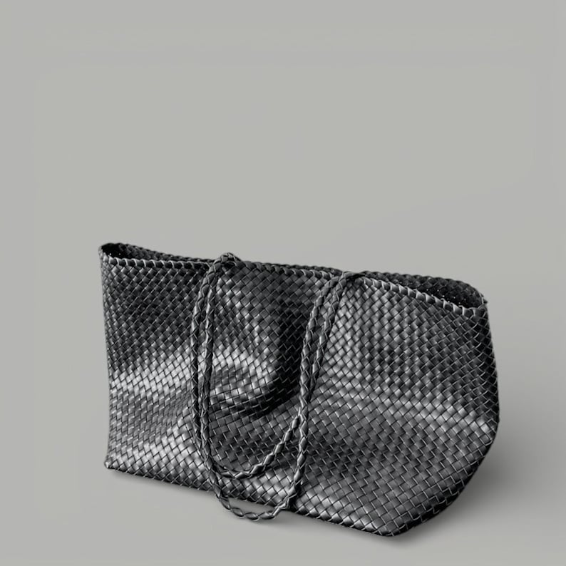 May include: A black tote bag with a woven design. The bag has two braided handles and a rectangular shape. The material appears to be leather or a leather-like material. The bag is set against a gray background.