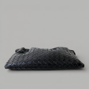 May include: A black woven leather clutch bag. The bag has a rectangular shape with a soft, gathered top. The leather is intricately woven in a basketweave pattern, creating a textured surface. The bag is set against a neutral background.