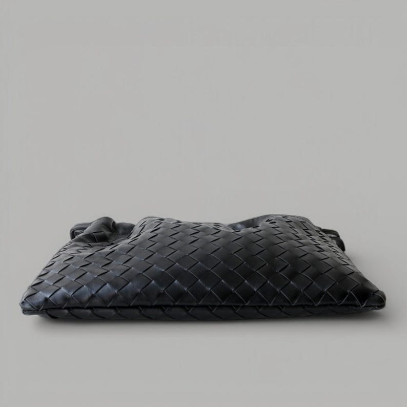 May include: A black woven leather clutch bag. The bag has a rectangular shape with a soft, gathered top. The leather is intricately woven in a basketweave pattern, creating a textured surface. The bag is set against a neutral background.