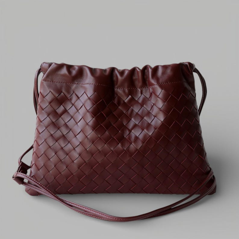 May include: A burgundy leather drawstring bag with a woven design. The bag features a long, thin strap for shoulder or crossbody use. The top of the bag is gathered with a drawstring closure.