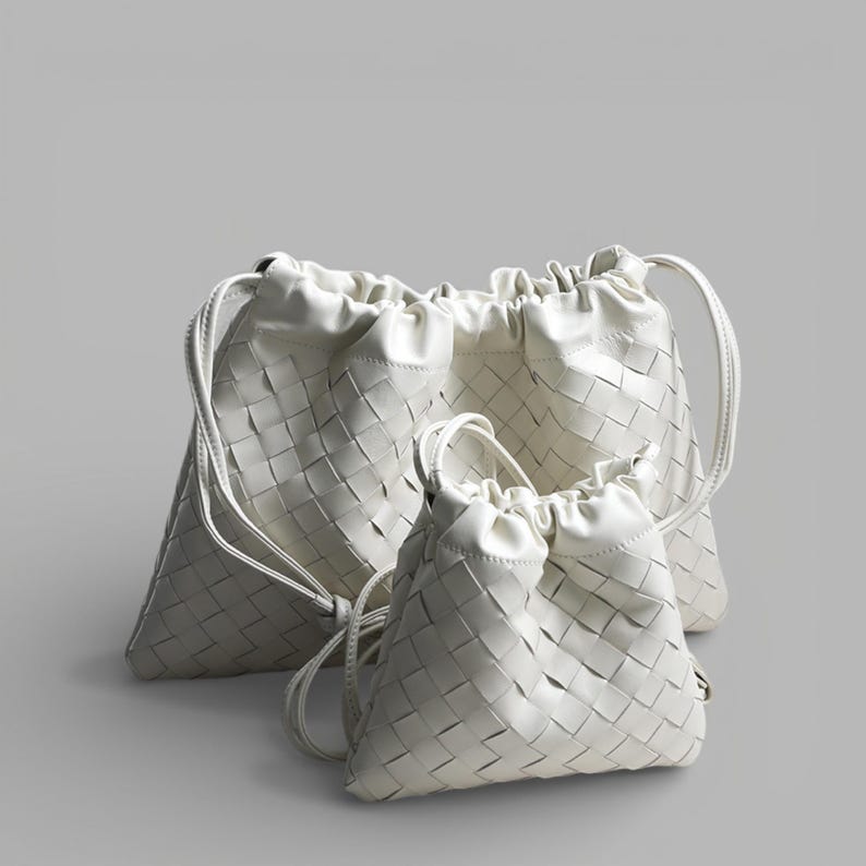 May include: Three white woven drawstring bags of different sizes are displayed against a grey backdrop. The bags feature a basket-weave pattern and a gathered top with a drawstring closure. The bags have a simple, elegant design.