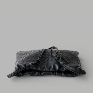 May include: Black woven leather clutch bag with drawstring closure. The bag has a textured, geometric pattern and a soft, gathered design. The drawstring is made of the same black leather.