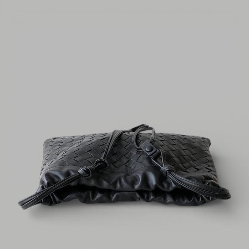 May include: Black woven leather clutch bag with drawstring closure. The bag has a textured, geometric pattern and a soft, gathered design. The drawstring is made of the same black leather.