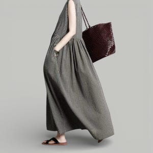 May include: A sleeveless, ankle-length dress in a black and white checkered pattern. The dress has a gathered waist and pockets. A dark brown woven tote bag is carried over the shoulder. The person is wearing black sandals.