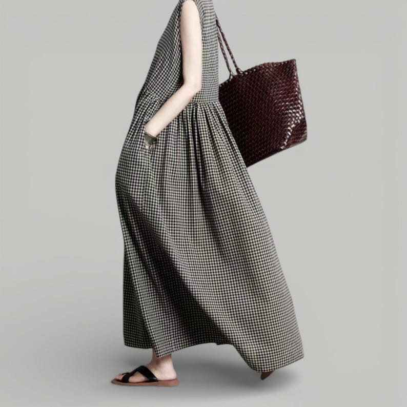 May include: A sleeveless, ankle-length dress in a black and white checkered pattern. The dress has a gathered waist and pockets. A dark brown woven tote bag is carried over the shoulder. The person is wearing black sandals.