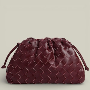 May include: A burgundy clutch with a woven pattern. The bag has a drawstring closure and is made of a soft material. The woven design is made of a darker burgundy color with a lighter burgundy color woven through it.