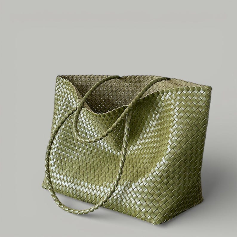 May include: A woven tote bag in a muted olive green hue. The bag features a rectangular shape with braided handles. The weave pattern is visible throughout the bag's exterior, creating a textured appearance. The bag is designed for carrying items.