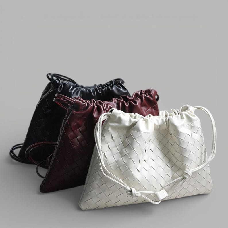 May include: Four woven leather drawstring bags in black, burgundy, and white. The bags have a basket weave pattern and a drawstring closure. The bags are displayed on a grey background.