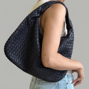 Pode incluir: Uma mala hobo preta com um padrão intrincado de pele entrançada em todo o exterior. A mala tem uma alça de ombro única integrada e uma silhueta descontraída e sem estrutura que se curva para se ajustar ao lado do corpo.