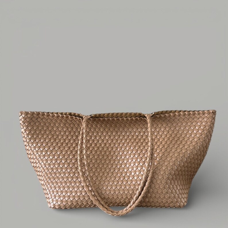 May include: A woven tote bag in a light brown color. The bag features a large, open design with two handles. The weave pattern is visible throughout the bag's exterior.