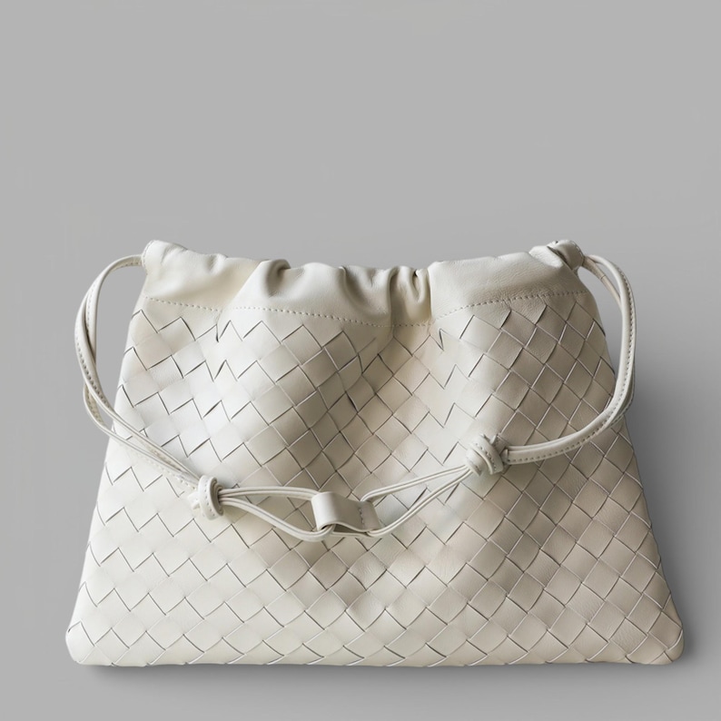 May include: Cream-coloured woven leather drawstring bag with a soft, gathered top. The bag has a diamond-patterned weave and a drawstring closure with knotted details. Curved handles are attached to the sides.
