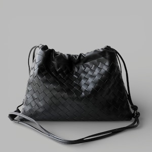 May include: A black leather shoulder bag with a woven design and drawstring closure. The bag has a long, thin strap for carrying. The woven pattern is a key feature. The bag is a neutral color and appears to be made of high-quality leather.