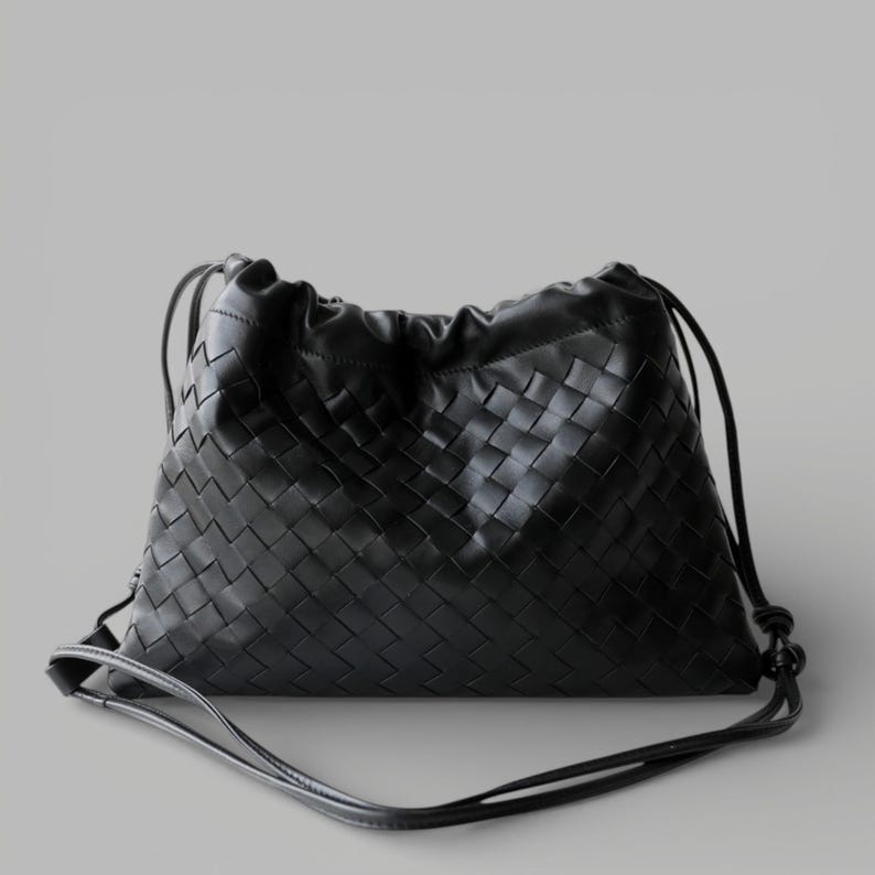May include: A black leather shoulder bag with a woven design and drawstring closure. The bag has a long, thin strap for carrying. The woven pattern is a key feature. The bag is a neutral color and appears to be made of high-quality leather.