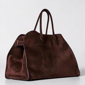 May include: A large, brown suede tote bag with two handles and a rectangular shape. The bag features a buckle closure on the side and a spacious interior. The bag is designed for carrying items and has a classic, elegant style.