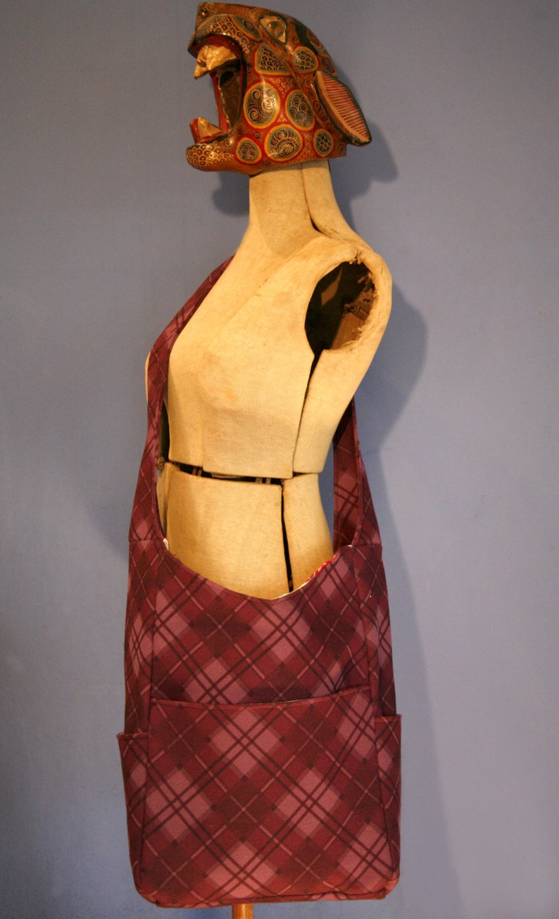 May include: A purple and gray plaid shoulder bag with a large pocket on the front. The bag is being modeled on a wooden mannequin.
