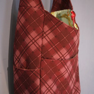 May include: A burgundy plaid tote bag with a single pocket on the front. The bag has a long strap and is made of a soft, durable fabric.