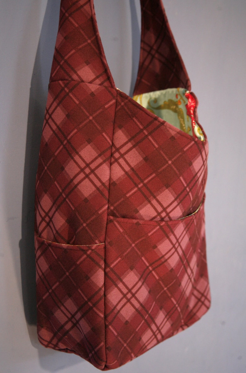 May include: A burgundy plaid tote bag with a single pocket on the front. The bag has a long strap and is made of a soft, durable fabric.
