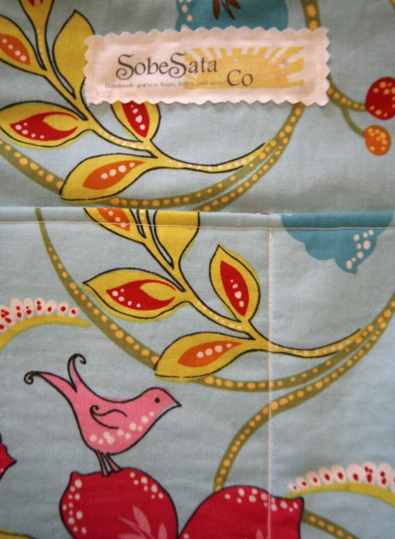 May include: A close-up of a blue and white fabric with a floral pattern. The fabric has a yellow, orange, and red floral design. The fabric has a white label with the text "SobeSata Co. Handmade purses, bags, totes, and more."