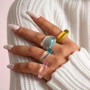 May include: A hand adorned with five colourful acrylic rings. The rings are in shades of blue, yellow, pink, and teal. The rings have a chunky, transparent design. The person is wearing a white sweater.