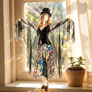 May include: A stained glass suncatcher depicting a figure wearing a black top hat, a black bodice, and a flowing skirt with multicolored, textured glass panels. The figure has long orange hair and outstretched arms with translucent black wings.
