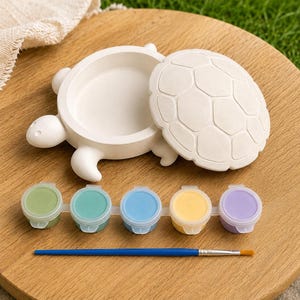 May include: A white turtle-shaped trinket box with a removable shell lid, accompanied by a strip of five small paint pots in green, teal, blue, yellow, and purple, and a single paintbrush with a blue handle and a golden-brown bristle tip.