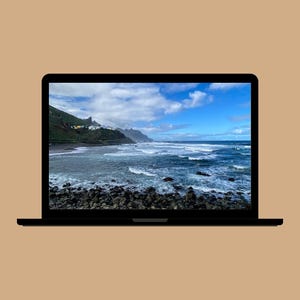 May include: A black laptop displays a scenic coastal view. The screen shows a blue ocean with white waves, a rocky shoreline, and a mountain range under a partly cloudy sky. The laptop is set against a tan background.