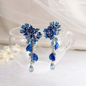 以下が含まれることがあります： 濃い青色のエナメル加工された花のクラスター、水色のパールビーズ、透明なクリスタルのアクセントが特徴のぶら下がりタイプのイヤリング。シルバー調のチェーンと、花のパーツから垂れ下がるティアドロップ型のチャームで構成されています。