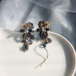 以下が含まれることがあります： エレガントなドロップイヤリングのペア。各イヤリングは、透明とゴールドのアクセント、濃い青のリボン、青とグレーの色合いのぶら下がりビーズを備えたフローラルデザインが特徴です。イヤリングは、金色の湾曲したワイヤーが付いています。