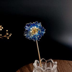 以下が含まれることがあります： パールとゴールドのアクセントが付いた、青と金のフラワーヘアピン。 花は半透明の素材でできており、金の茎が付いています。 ヘアアクセサリーは、暗い背景に対して透明なガラスの花瓶に飾られています。