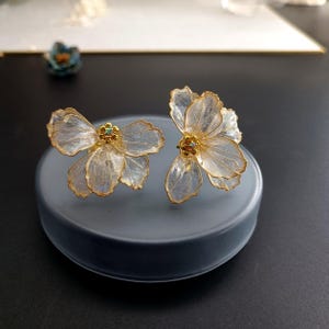 以下が含まれることがあります： 花型のイヤリングのペア。各イヤリングは、縁を縁取る金色のワイヤーが付いた、繊細で半透明の花びらを特徴としています。各花の中心は、小さくカラフルな宝石で飾られています。イヤリングは灰色の表面に表示されています。